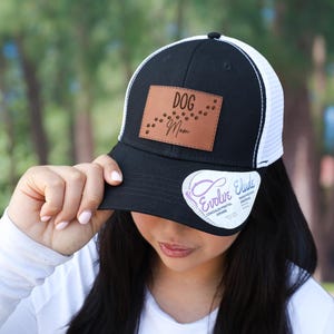 May include: Black and white trucker hat with a mesh back. The hat features a brown leather patch with the words "Dog Mom" and paw prints. A white sticker with the words "Evolve Elude" is on the brim.
