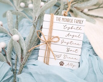 May include: White wooden ornament with the text "The Monroe Family" and a list of names: Elizabeth, Angel, Justine, Brit, Diana, and Ally. The ornament is decorated with a brown twine bow and a small paw print.