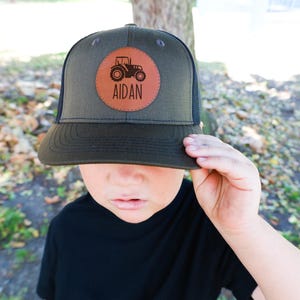 May include: A green and black trucker hat with a brown leather patch featuring a tractor and the name "AIDAN".