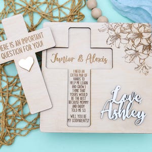 May include: Wooden cross with the text "There is an important question for you!" and a small heart. A larger wooden cross with the text "Junior & Alexis" and the text "I need an extra pair of hands to help me learn and grow I think that yours would be the best because mommy and daddy told me so Will you be my godparents?" A white wooden cutout of the text "Love Ashley" is on the right side of the cross.