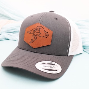May include: Gray and white trucker hat with a brown leather patch. The patch features a fist bump design with the words "FATHER", "DAD", and "MASON". The hat has a curved brim and a snapback closure.