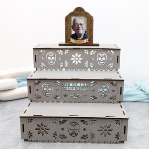 May include: A three-tiered light gray wooden display stand featuring laser-cut sugar skull and floral patterns. The middle tier displays the text "DIA DE LOS MUERTOS". A small wooden frame sits on top, holding a photograph of an older man with the word "GRANDPA" engraved on a banner below the image.