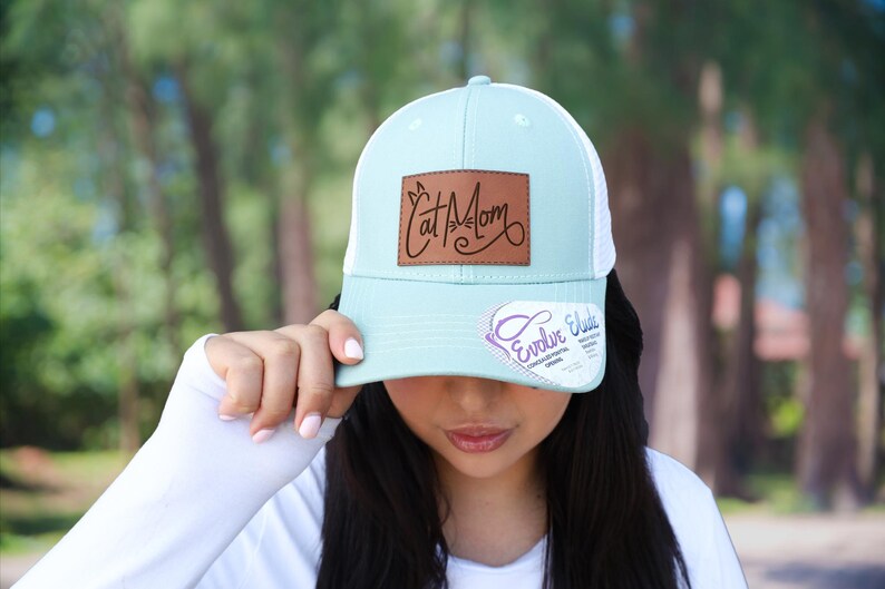 May include: A light blue and white trucker hat with a brown leather patch that reads "Cat Mom" in a script font. The hat has a curved brim and a mesh back. The hat also has a small tag with the text "Evelyn Elude".