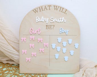 May include: Wooden gender reveal board with the text "WHAT WILL Baby Smith BE?" in a script font. The board is divided into two sections, one with pink bows for "Girl" and the other with blue onesies for "Boy".