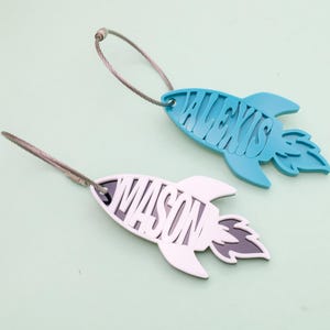 May include: Two rocket-shaped luggage tags. One is light blue with the name "ALEXIS" cut out, and the other is white with "MASON" cut out. Both have a silver-colored wire loop for attachment.