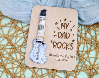 May include: A wooden Father's Day card with a guitar-shaped cutout holding a rolled-up dollar bill. The card reads "MY DAD ROCKS" with stars and lightning bolts, and "Happy Father's Day Dad! Love, Jacob" below.