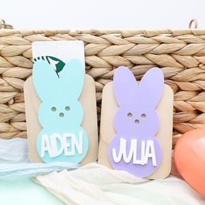 May include: Two Easter bunny-shaped gift card holders, one light blue and one purple, each with a name in white. The holders are on wooden blocks, with a woven basket in the background. Easter eggs in orange, yellow, and pink are also visible.