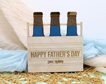 May include: A wooden beer bottle holder with three blue and brown beer bottle cutouts. The holder is engraved with "HAPPY FATHER'S DAY" and "Love, Ashley". The background includes blue and white fabric.
