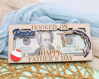 May include: Wooden Father's Day gift with the words "Hooked on Papa" and "Happy Father's Day" cut out. A fishing bobber and a fishing hook are attached to the wooden frame. The frame is designed to hold a US twenty-dollar bill.