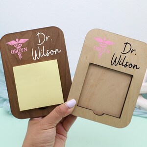 May include: Two wooden memo holders, one dark brown and one light brown, with rounded corners. Both have the text "Dr. Wilson" and a pink OBGYN symbol. The dark brown holder has a yellow sticky note pad.