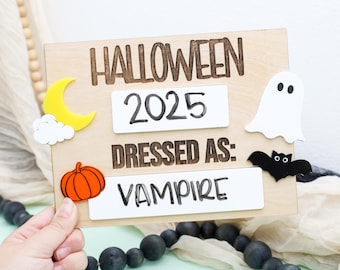 May include: Wooden Halloween sign with the text "HALLOWEEN 2025 DRESSED AS: VAMPIRE". Includes decorative cutouts of a yellow moon, white cloud, orange pumpkin, white ghost, and black bat. The sign is held by a hand.