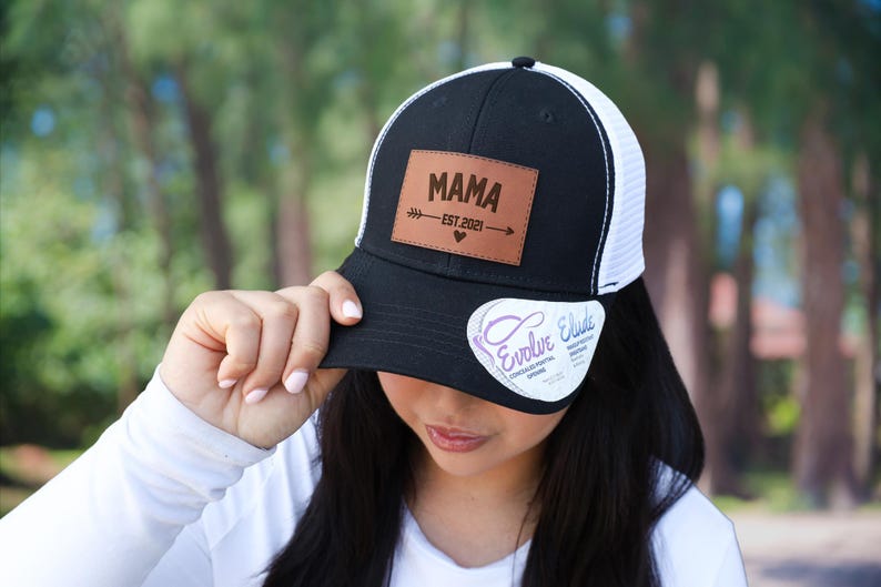 Pu&ograve; includere: Cappellino da camionista nero e bianco con una toppa in pelle che recita "MAMA EST. 2021" con un design a freccia e cuore. Il cappellino ha una visiera curva e un retro in rete.