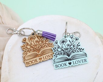 May include: Two wooden book-shaped keychains with floral designs and the text "BOOK LOVE" on a white surface. One keychain is light brown with purple tassel and the other is light blue.