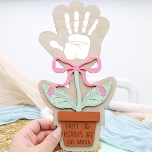 May include: A wooden handprint flower craft with a pink bow and green leaves. The flower is in a brown pot with the text "Happy First Mother's Day Love, Isabella".