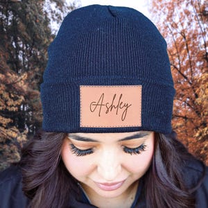 May include: A black beanie with a brown leather patch that says "Ashley" on it. The beanie is worn by a person with long dark hair.