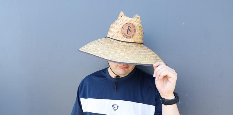 Pode incluir: Uma pessoa usando um chap&eacute;u de palha com um remendo de couro marrom que diz "S&C". O chap&eacute;u tem uma aba larga e est&aacute; puxado sobre os olhos da pessoa. A pessoa est&aacute; usando uma camisa listrada azul e branca.