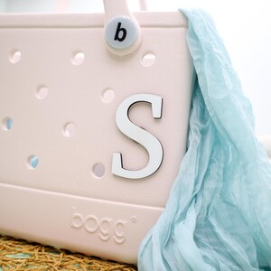 May include: A pink and white tote bag with a white letter "S" on the front. The bag has a blue fabric draped over it and the word "bogg" is printed on the bottom.
