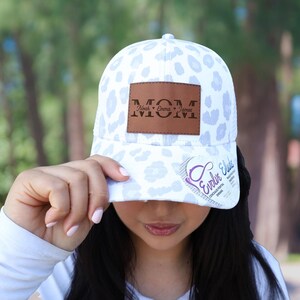 May include: White baseball cap with a light blue leopard print pattern. The cap features a brown leather patch with the word "MOM" and the names "Noah, Emma, James" engraved. The brim has a sticker with the text "Evolin Elise".