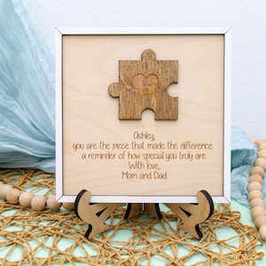 May include: A white wooden frame with a light brown background. The frame has a cutout of a puzzle piece with a heart in the center. The text "Ashley you are the piece that made the difference a reminder of how special you truly are With love, Mom and Dad" is engraved on the frame.
