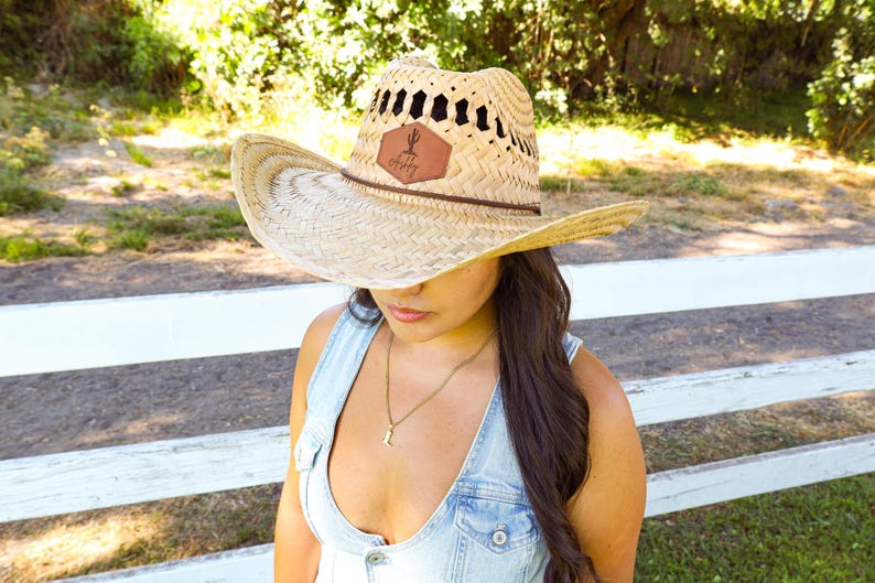 May include: A woman wearing a straw cowboy hat with a leather patch that says "Happy" on it. She is wearing a denim overall dress and has long brown hair.