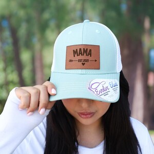 Pu&ograve; includere: Cappellino da camionista azzurro e bianco con una toppa in pelle marrone con la scritta "MAMA EST 2021" con un design a freccia e cuore. Il cappello ha una visiera curva e un retro in rete.