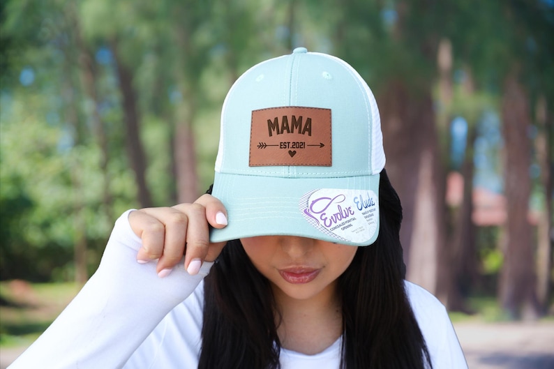 Pu&ograve; includere: Cappellino da camionista azzurro e bianco con una toppa in pelle marrone con la scritta "MAMA EST 2021" con un design a freccia e cuore. Il cappello ha una visiera curva e un retro in rete.
