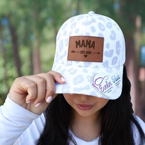 Pu&ograve; includere: Cappellino da baseball bianco con un motivo leopardato lilla chiaro. Il cappellino presenta una toppa in pelle marrone con la scritta "MAMA" e il testo "EST. 2021". Il cappellino ha anche il testo "Evolve Elude" sulla tesa.