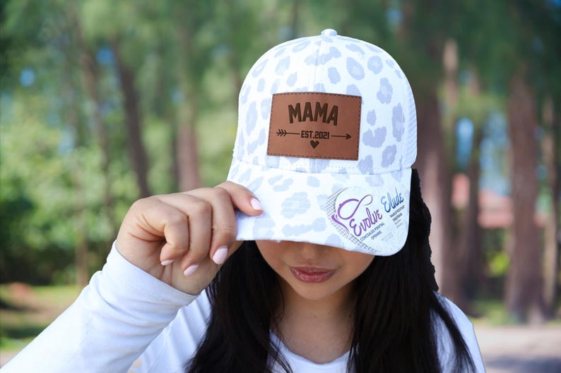 Pu&ograve; includere: Cappellino da baseball bianco con un motivo leopardato lilla chiaro. Il cappellino presenta una toppa in pelle marrone con la scritta "MAMA" e il testo "EST. 2021". Il cappellino ha anche il testo "Evolve Elude" sulla tesa.
