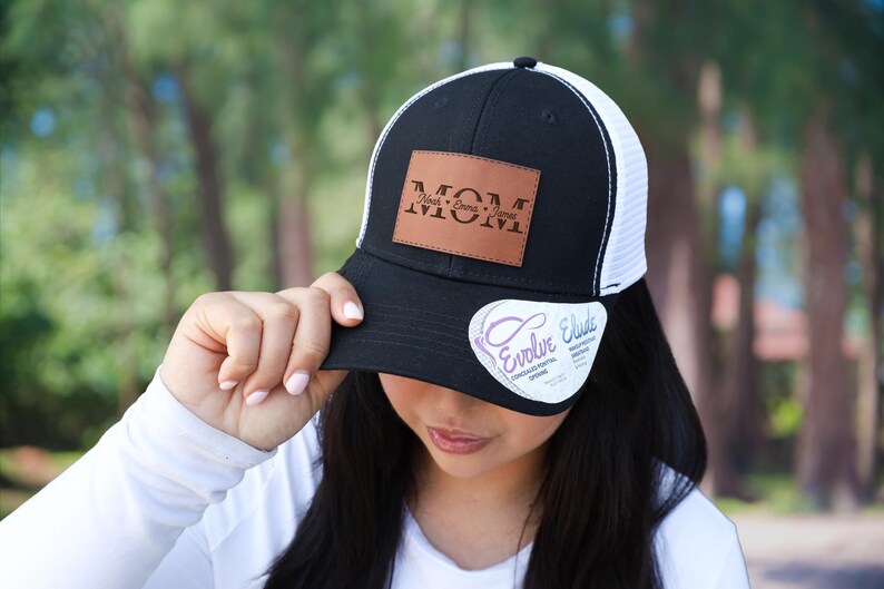 May include: Black and white trucker hat with a brown leather patch that reads "MOM". The hat has a curved brim and a white mesh back. A white sticker with the text "Evolve Elude" is on the brim.