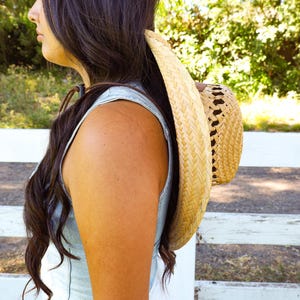 May include: A woman with long brown hair wearing a light blue sleeveless top and a large straw hat.