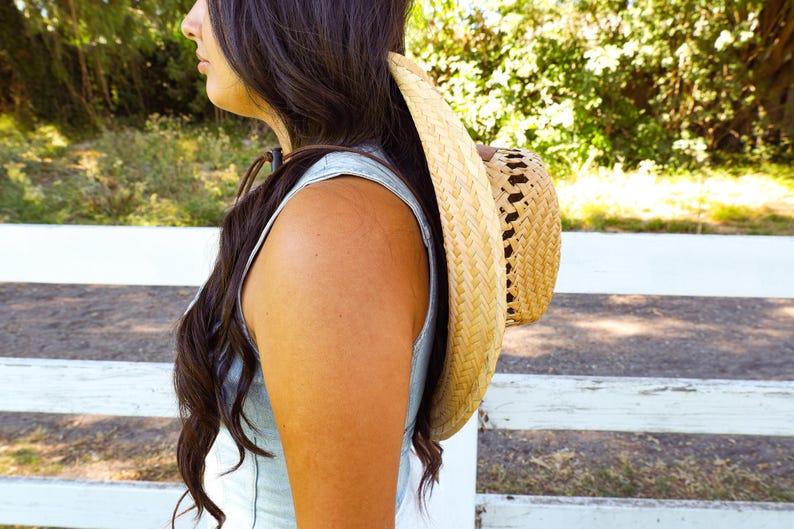 May include: A woman with long brown hair wearing a light blue sleeveless top and a large straw hat.