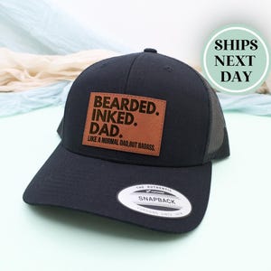 May include: A black snapback hat with a mesh back and a curved bill. The hat features a brown leather patch with the words "BEARDED. INKED. DAD." in white lettering. Below the text is the phrase "LIKE A NORMAL DAD, BUT BADASS."