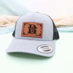 May include: A gray trucker hat with a black mesh back and a curved brim. A brown leather patch on the front features the letter "D" and the name "Brandon" in a decorative font. The hat has a snapback closure.