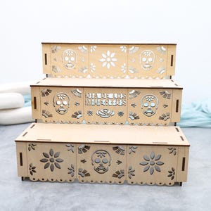 May include: Three-tiered wooden display stand with laser-cut Day of the Dead themed designs. The stand features cut-out skulls, flowers, and the text "Dia de los Muertos". The natural wood color provides a neutral backdrop.