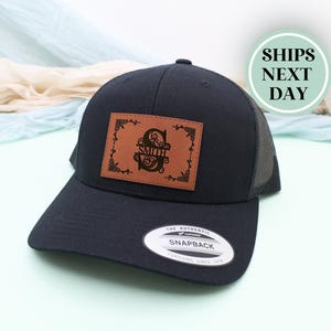 May include: A navy blue trucker hat with a brown leather patch featuring the name "Smith" in a decorative font. The hat has a mesh back and a curved brim. The hat has a Snapback tag.