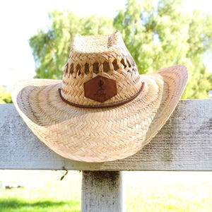 May include: A straw cowboy hat with a brown leather patch that says "Get Dirty" hanging on a white wooden fence.