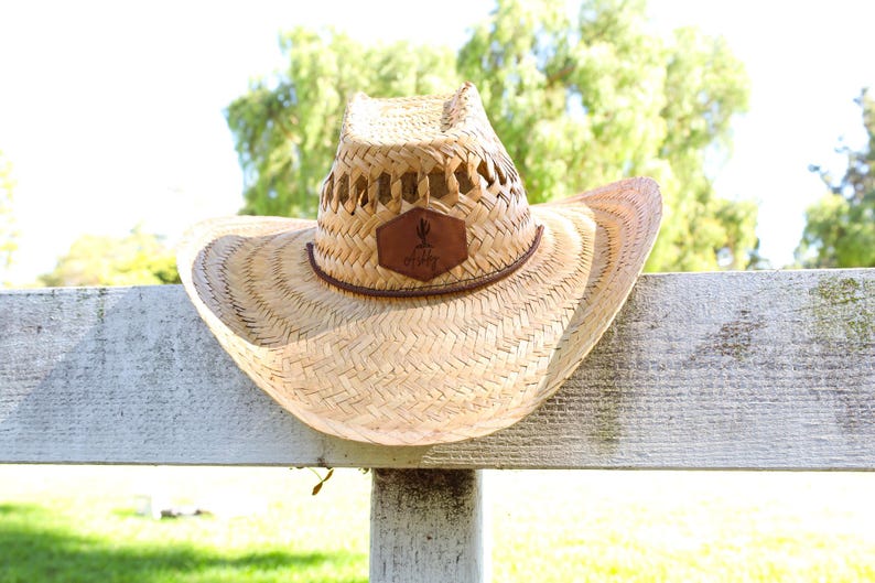 May include: A straw cowboy hat with a brown leather patch that says "Get Dirty" hanging on a white wooden fence.