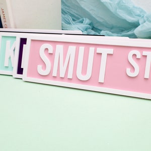 May include: A set of five pink, purple, black, and light blue street signs with white lettering that reads "SMUT ST".
