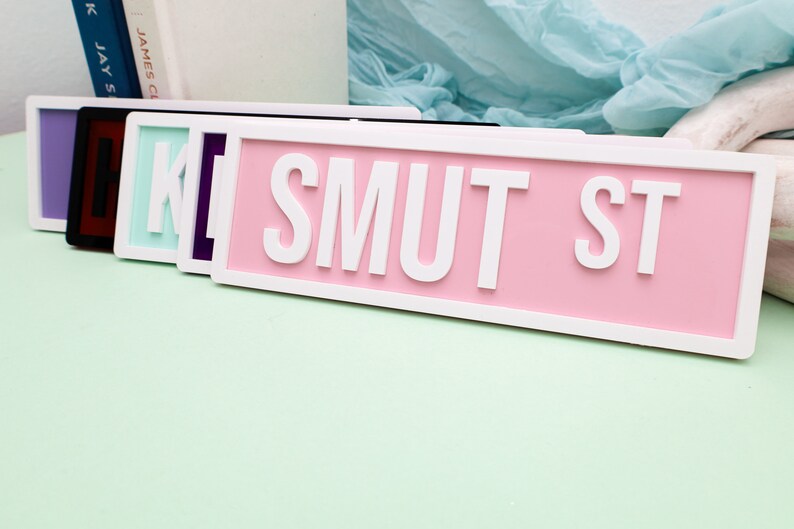 May include: A set of five pink, purple, black, and light blue street signs with white lettering that reads "SMUT ST".