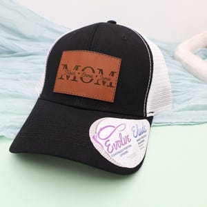 May include: Black and white trucker hat with a brown leather patch that reads "MOM" with the names Noah, Emma, and James. The hat has a curved brim and a white mesh back. A sticker with the text "Evolve Elude" is on the brim.