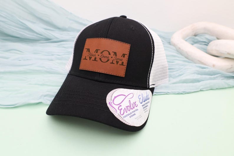 May include: Black and white trucker hat with a brown leather patch that reads "MOM" with the names Noah, Emma, and James. The hat has a curved brim and a white mesh back. A sticker with the text "Evolve Elude" is on the brim.