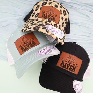 May include: Three trucker hats with the text "Life is better at the River" on a brown leather patch. One hat is black, one is light blue, and one has a leopard print pattern. Each hat has a white mesh back.