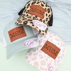 May include: Three baseball caps with the word "MOM" on a brown leather patch. One cap has a leopard print, another is light blue, and the third is white with a gray pattern. Each cap has a mesh back and a curved brim.