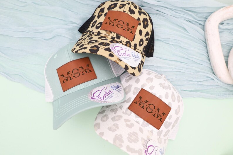 May include: Three baseball caps with the word "MOM" on a brown leather patch. One cap has a leopard print, another is light blue, and the third is white with a gray pattern. Each cap has a mesh back and a curved brim.