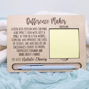 May include: Wooden sign with the text "Difference Maker" and a definition of the word. The sign has a space for a sticky note and a pen holder.