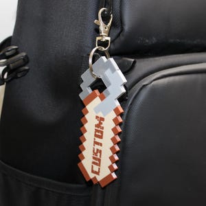 May include: A pixelated keychain with the word "CUSTOM" in a brown and white design, attached to a black backpack. The keychain is made of a gray and brown material and is attached with a silver clasp.