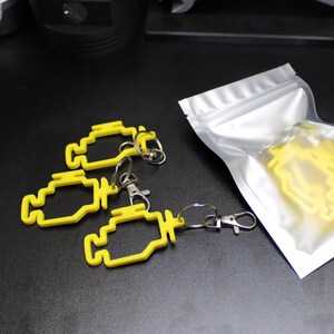 May include: Yellow engine-shaped keychains with silver key rings and clasps. Some keychains are loose, while others are in a clear, sealed bag. The keychains are on a black surface.