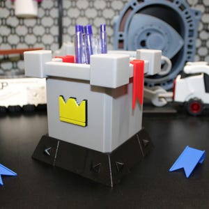 May include: A gray castle-shaped desk organizer with a yellow crown emblem, red flags, and blue accents. The organizer holds pens and pencils. Two blue pennants are in front of the organizer. Other toy objects are in the background.