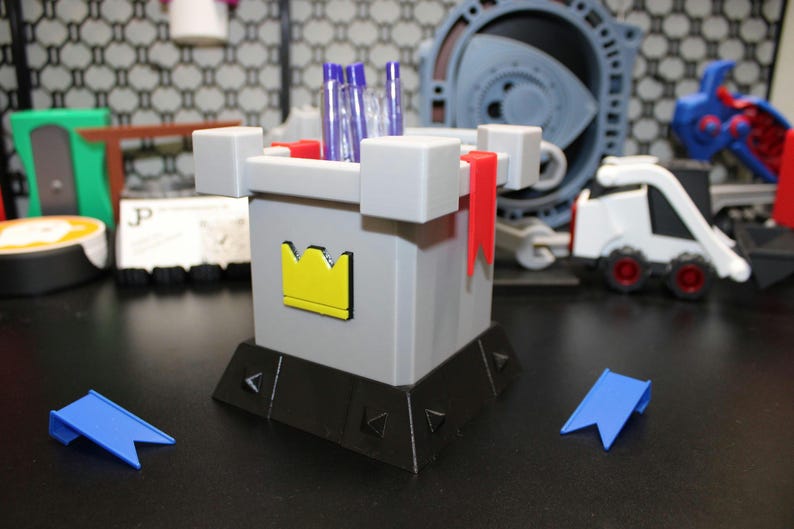 May include: A grey castle-shaped desk organiser with a yellow crown emblem, red flags, and blue accents. The organiser holds pens and pencils. Two blue pennants are in front of the organiser. Other toy objects are in the background.