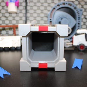 May include: A grey, rectangular toy structure with red accents and a hollow interior. Two blue, flag-shaped pieces are in front of the structure. Other toys are in the background, including a white vehicle with red wheels.